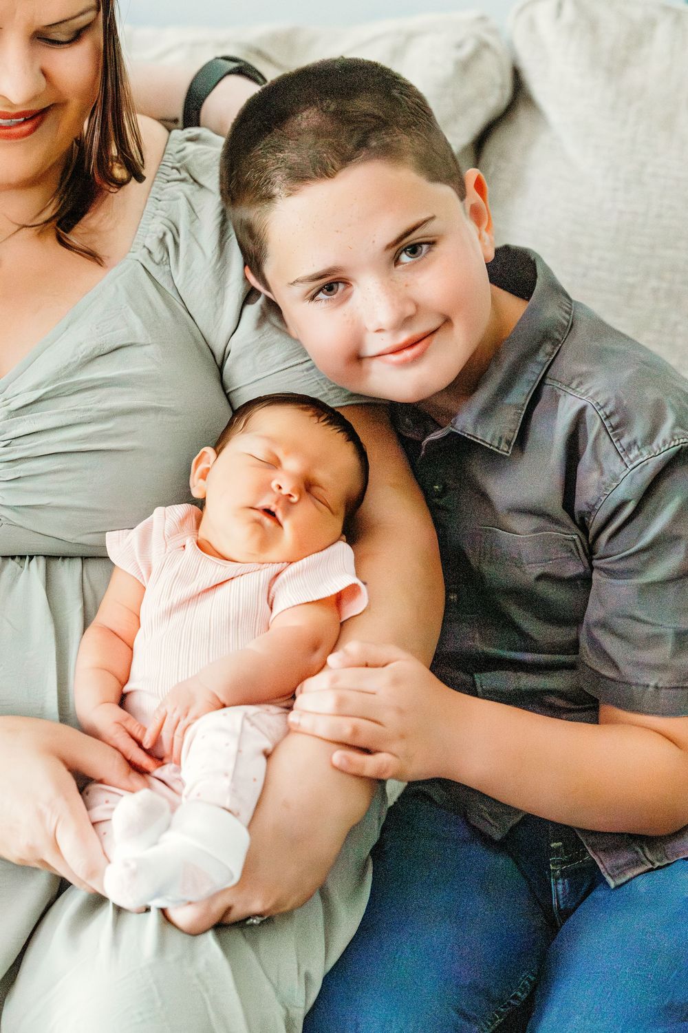 A smiling boy holds a sleeping newborn baby in pink while resting against a woman in a sage green dress.