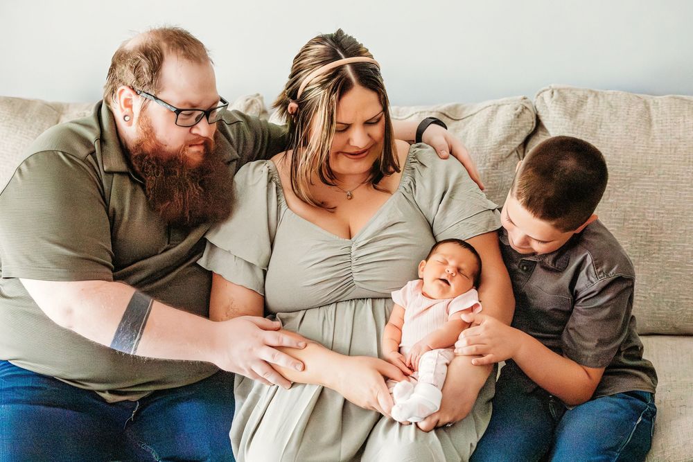 Family of four sitting on a couch, parents smiling at newborn baby held by mother, with young boy beside them.