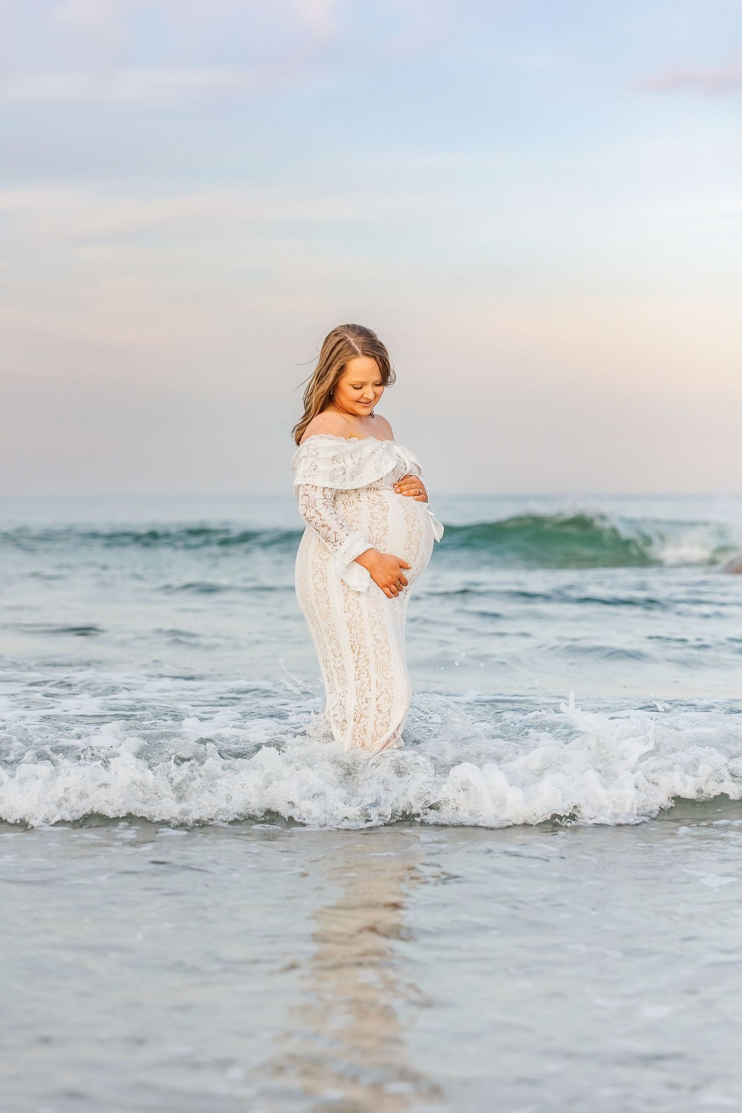Pregnant woman in white lace dress standing in ocean waves on beach at sunset, cradling her baby bump.