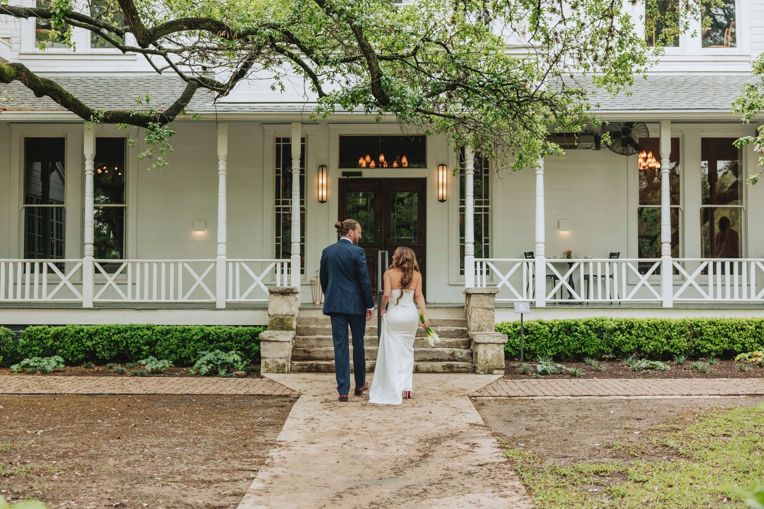 Bride and groom walking toward elegant white Southern plantation wedding venue with wraparound porch.