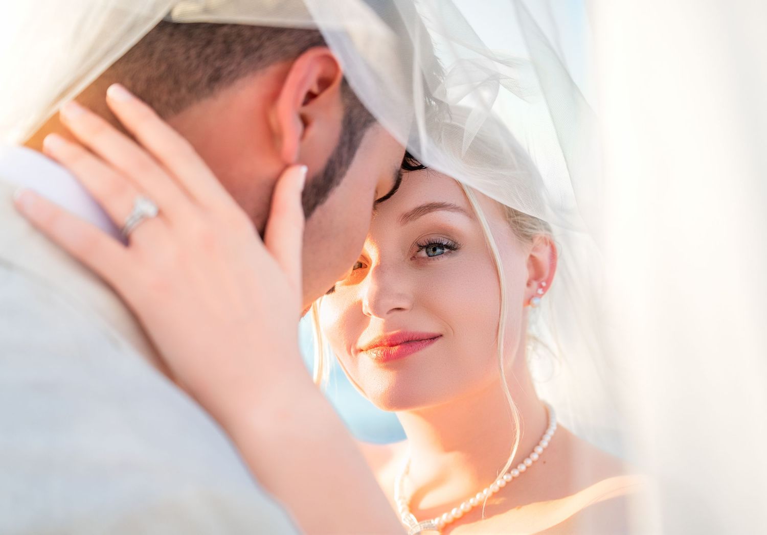 Bride and groom share an intimate moment under a wedding veil, with the bride wearing a pearl necklace and diamond ring.