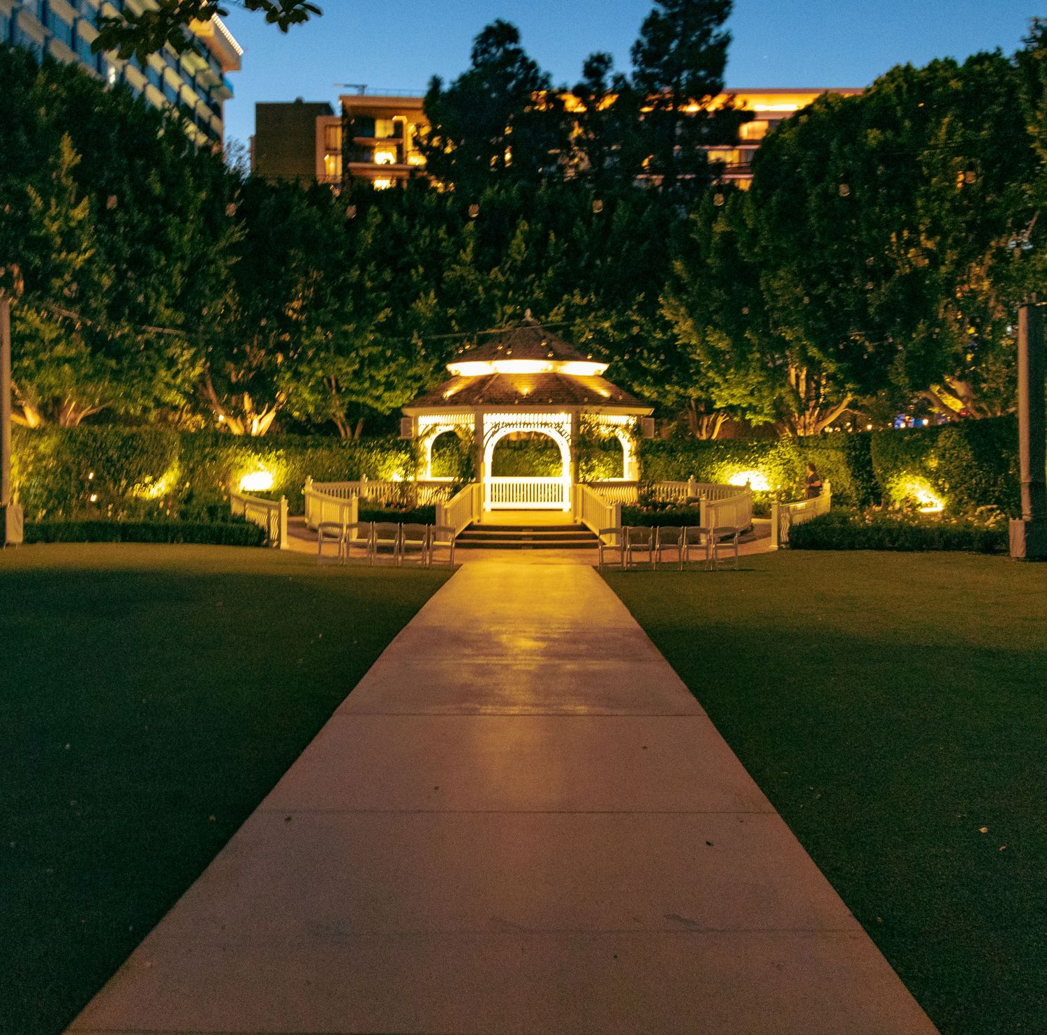 Illuminated gazebo at dusk along a lit garden pathway surrounded by lush trees and manicured lawn.