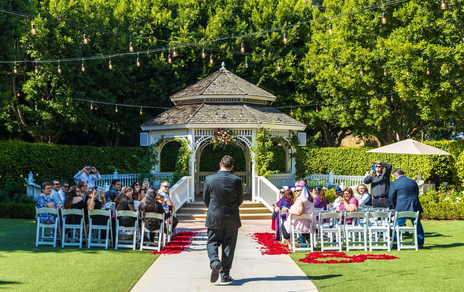 Outdoor wedding ceremony at a gazebo venue with white chairs, red aisle, and string lights on a sunny day.
