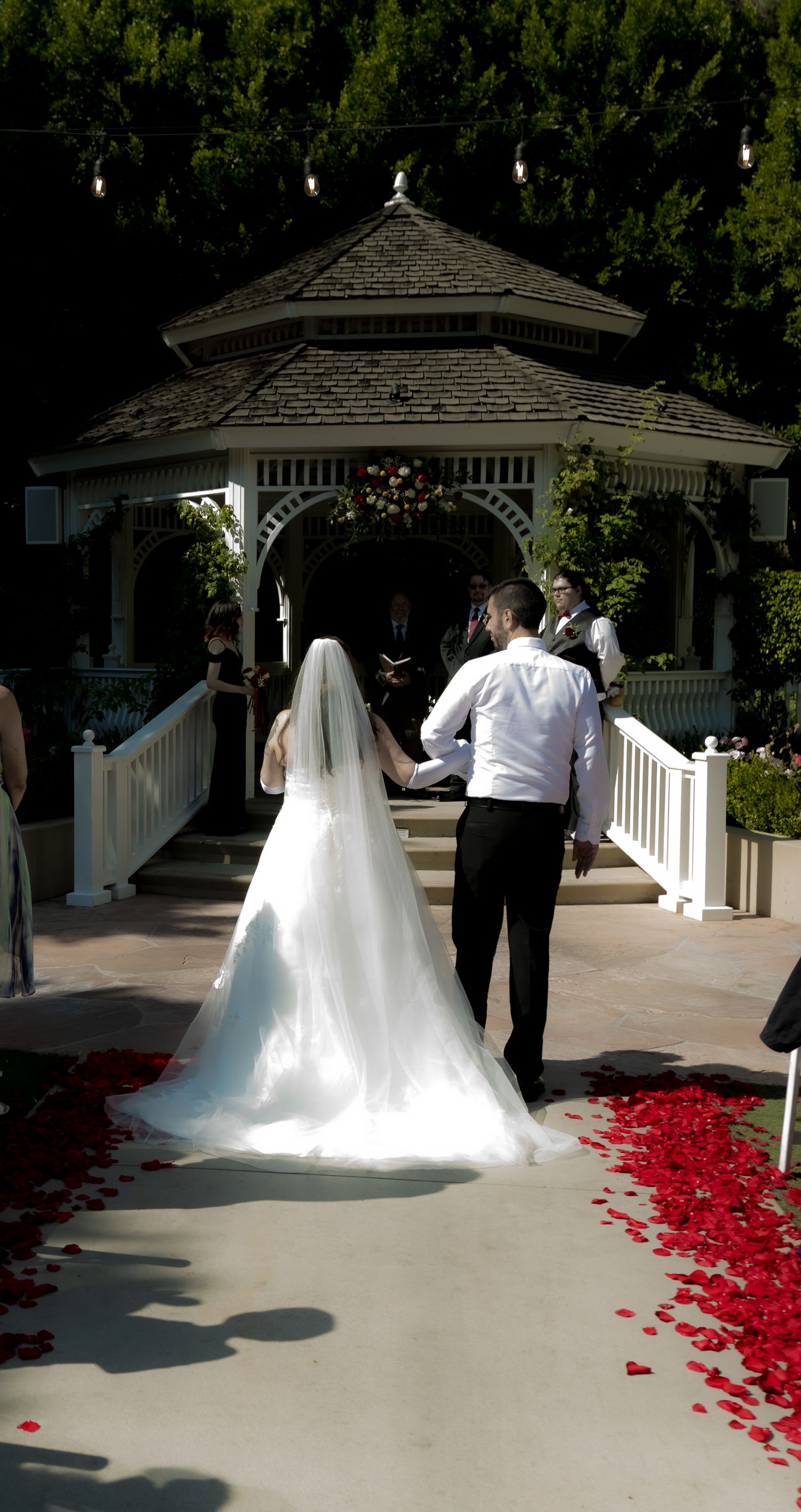 Bride in white gown and veil walks with groom toward floral gazebo, red rose petals lining the wedding aisle.