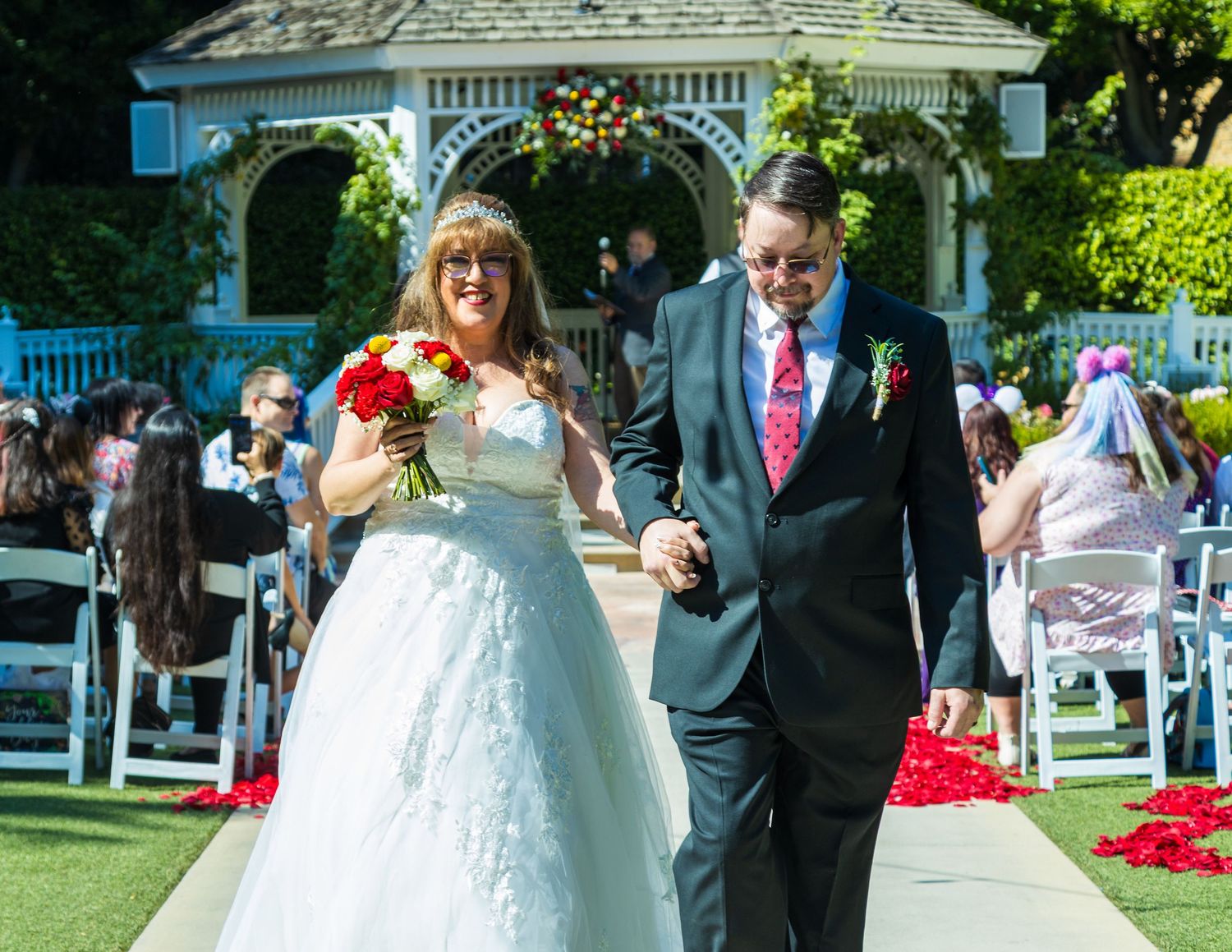 Bride and groom walk down rose petal aisle after outdoor wedding ceremony near white gazebo.