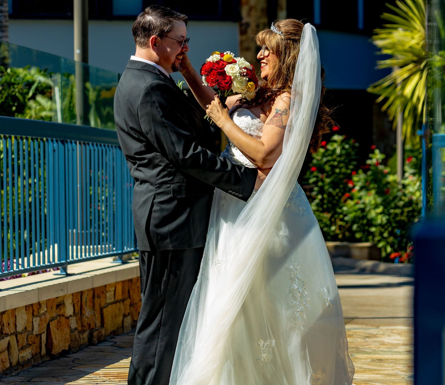 Bride and groom share a romantic moment outdoors at a tropical wedding venue, bride holding a red bouquet.