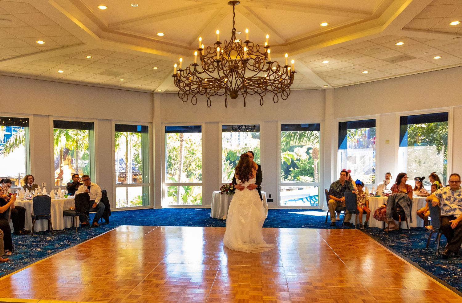 Elegant wedding reception first dance under a chandelier in a bright ballroom with large windows and seated guests.