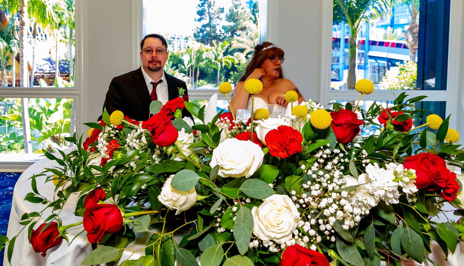 Bride and groom seated behind elegant table with red and white rose floral arrangements at wedding reception.
