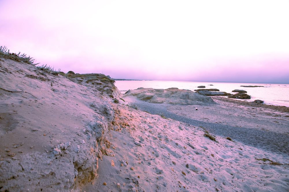 Sandy beach dunes at sunset with a calm sea and pink-purple sky.