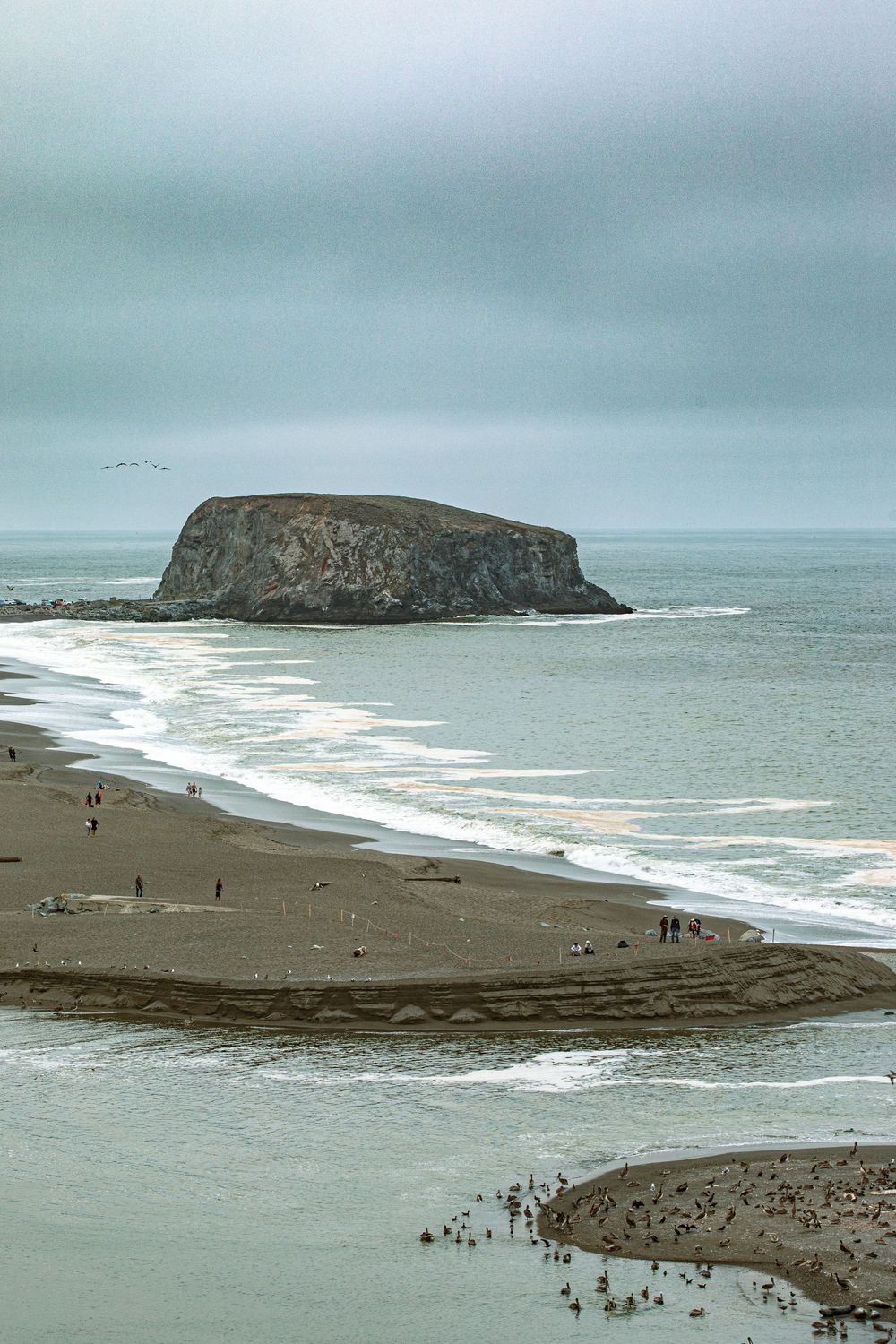 Overcast coastal scene with a rocky sea stack offshore, sandy beach with scattered visitors, and calm ocean waves.