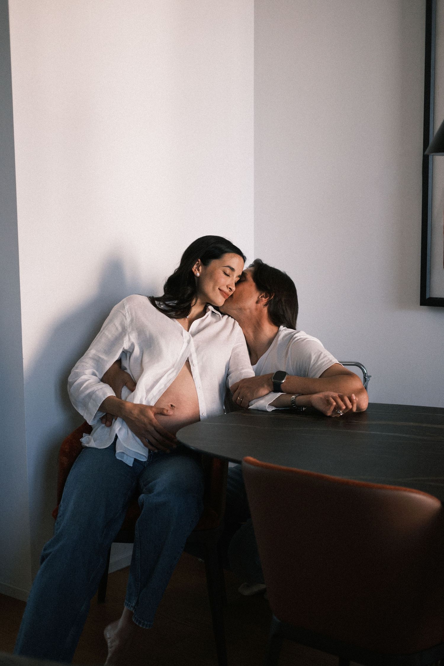 A pregnant woman in a white shirt receives a tender kiss from her partner while sitting together at a dark round table.