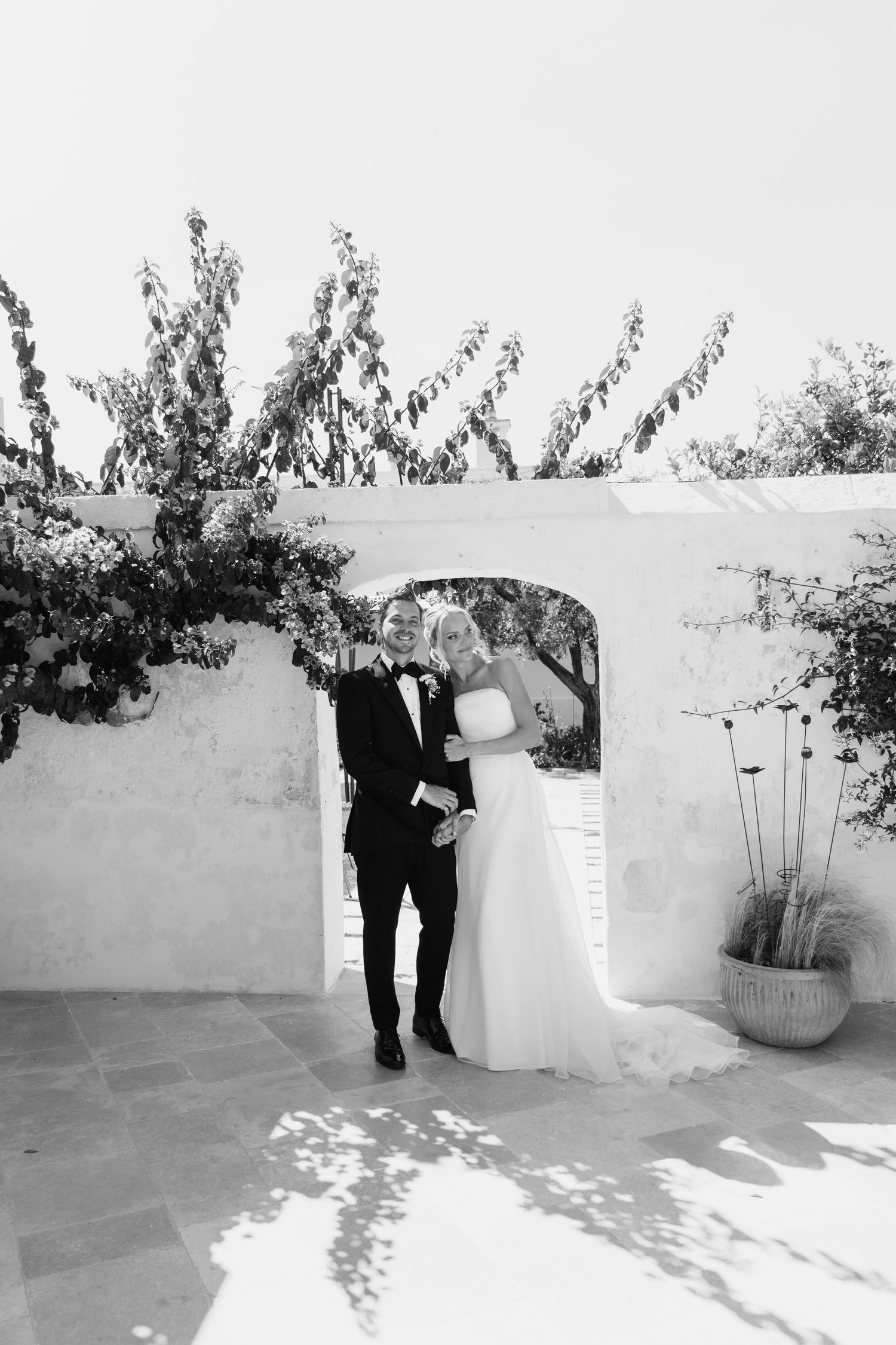 A bride and groom walk hand in hand through a sunlit garden path, smiling at each other on their wedding day.