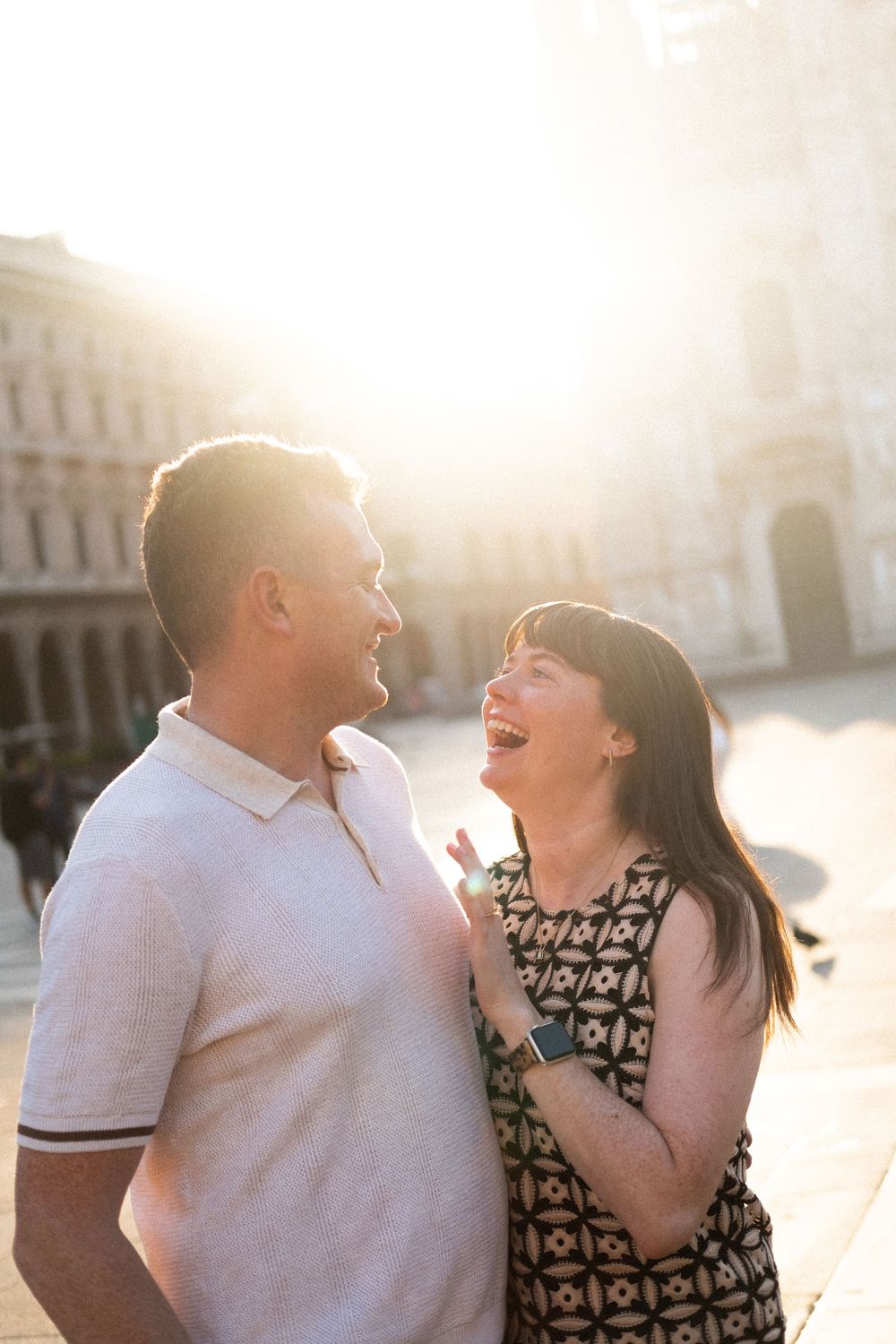 A joyful couple laughing together in a sunlit European plaza, woman in patterned dress, man in white polo shirt.