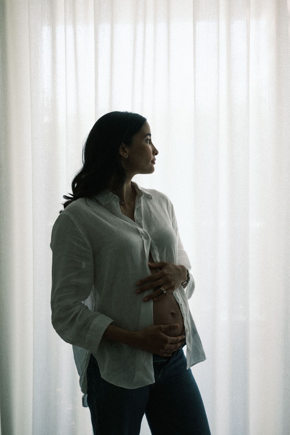 Pregnant woman in white shirt cradling her baby bump while standing by sheer curtains in soft natural light.