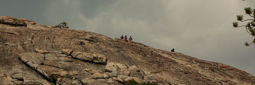 Hikers climbing a massive granite rock face under a cloudy sky in a scenic mountain landscape.