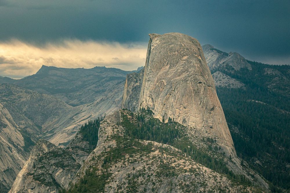 Dramatic view of Half Dome in Yosemite National Park under stormy skies, surrounded by granite cliffs and pine forests.