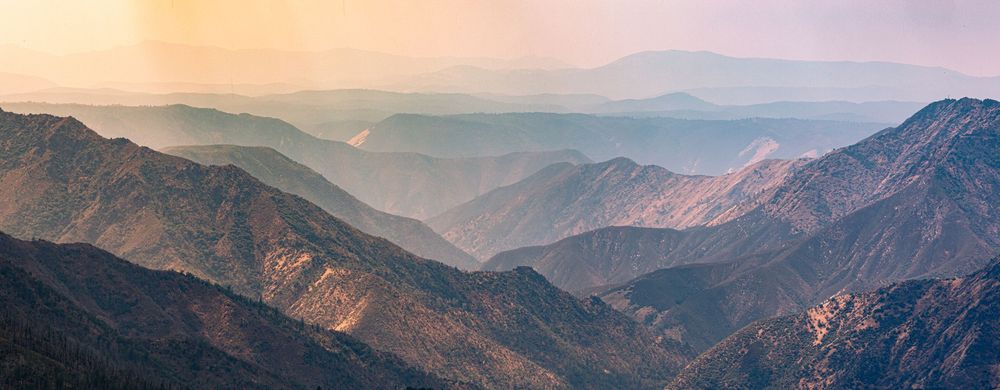 Panoramic sunset view of layered mountain ranges with warm hazy golden light casting soft shadows over rugged peaks.