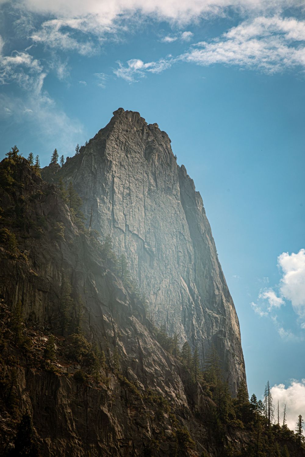 Towering granite cliff face with pine trees rising against a blue sky with scattered clouds in a mountain landscape.