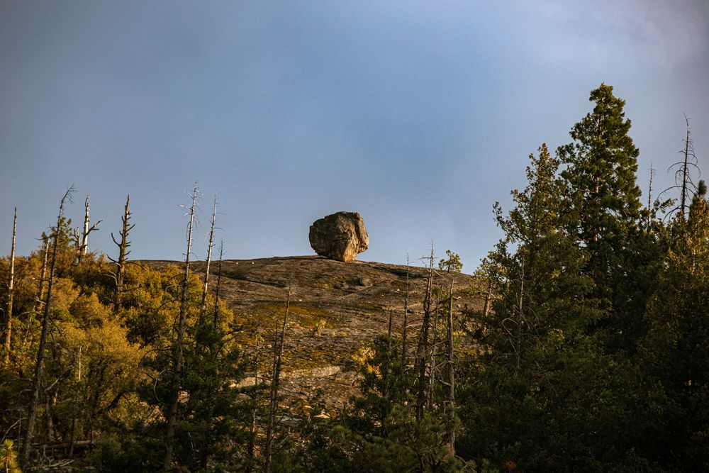A large balanced rock perches atop a granite hillside surrounded by pine trees under a blue sky.
