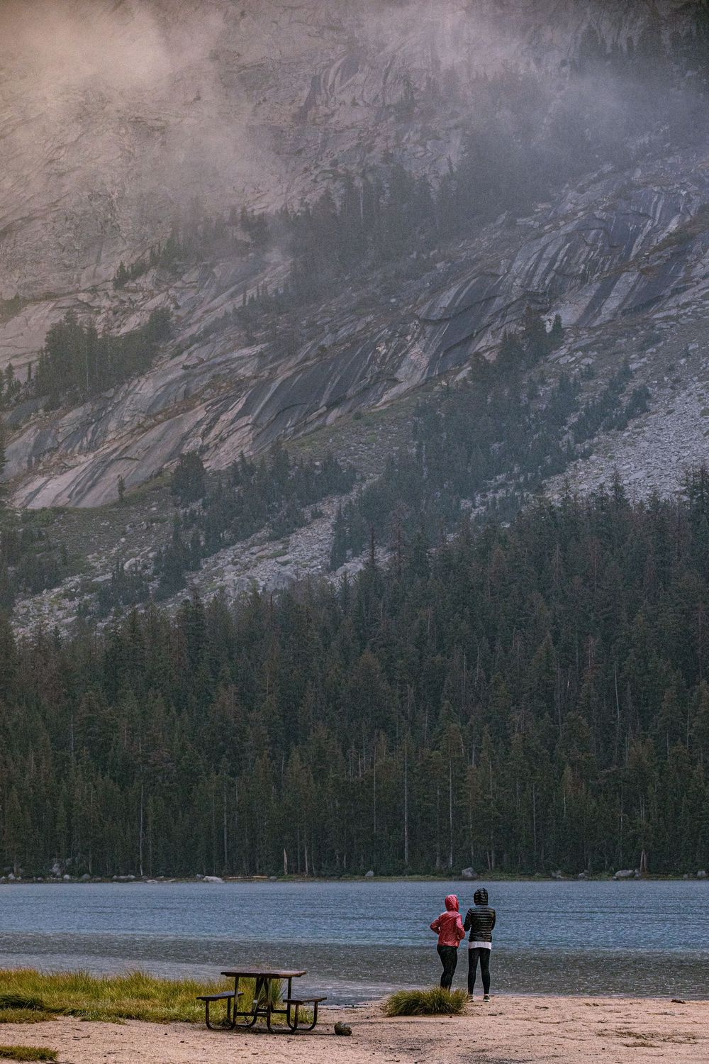 Two people stand by a mountain lake with pine trees and rocky cliffs in misty light.