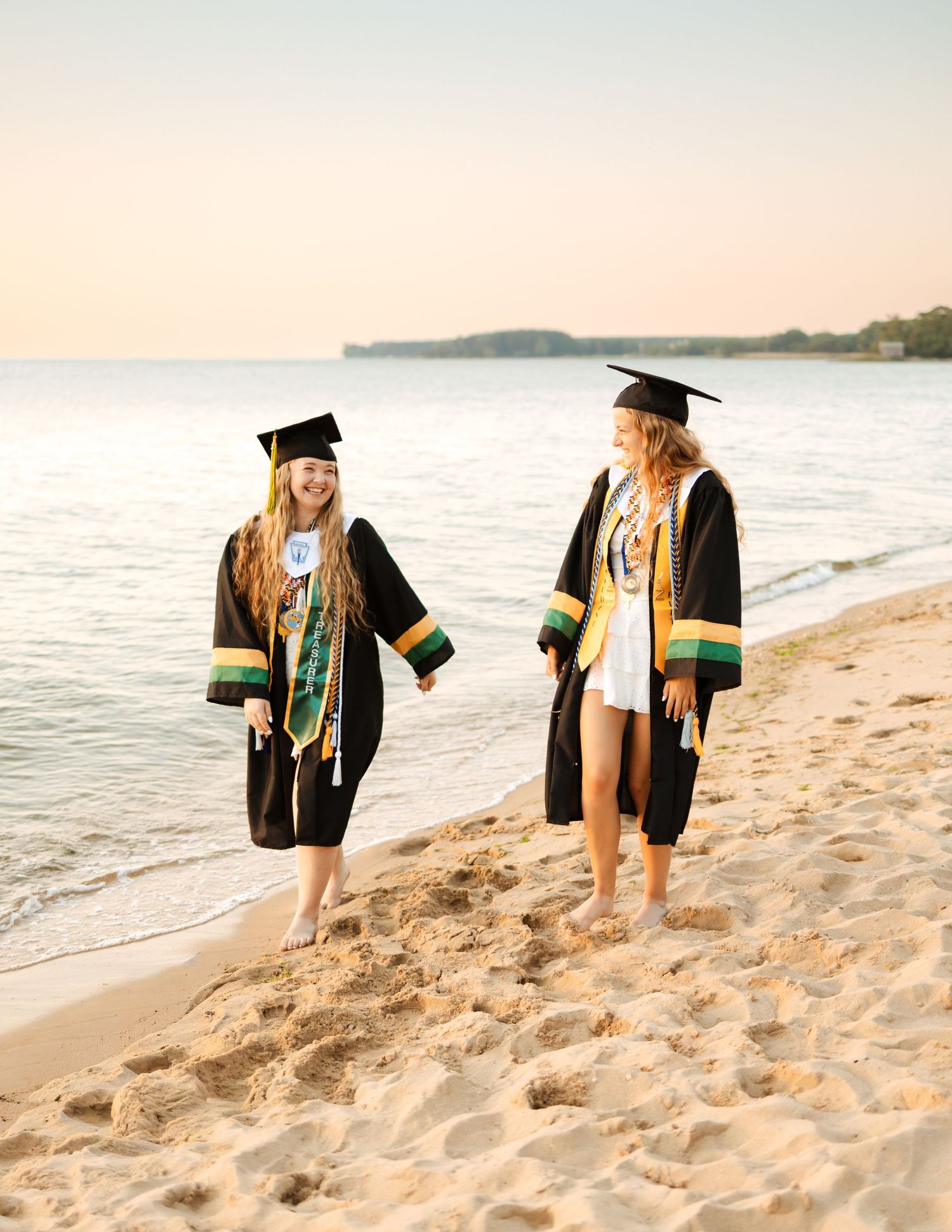 Two female graduates in black caps and gowns walk barefoot on a sandy beach at sunset.