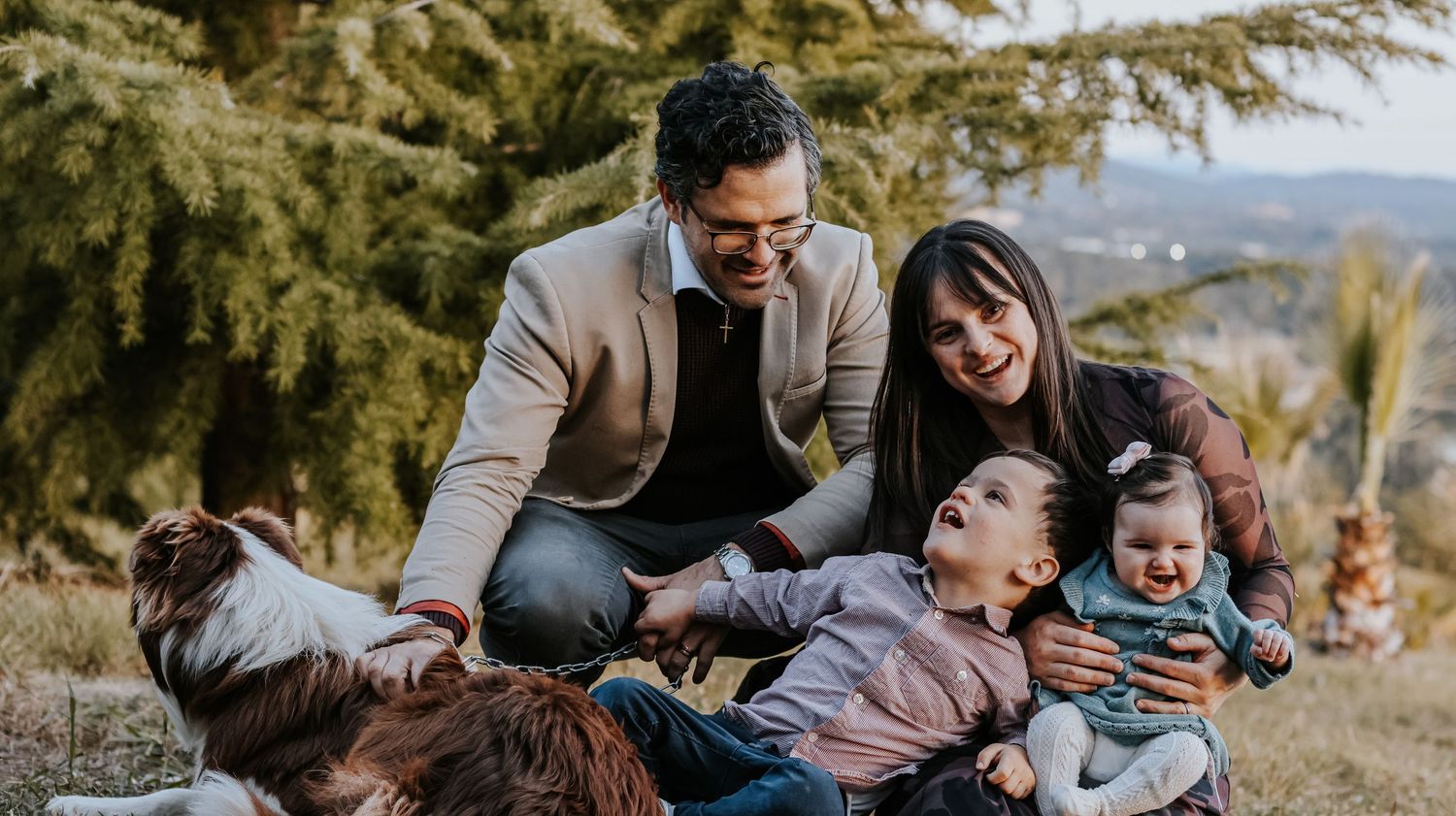 Happy family with two young children laughing together outdoors near pine trees on a sunny day.