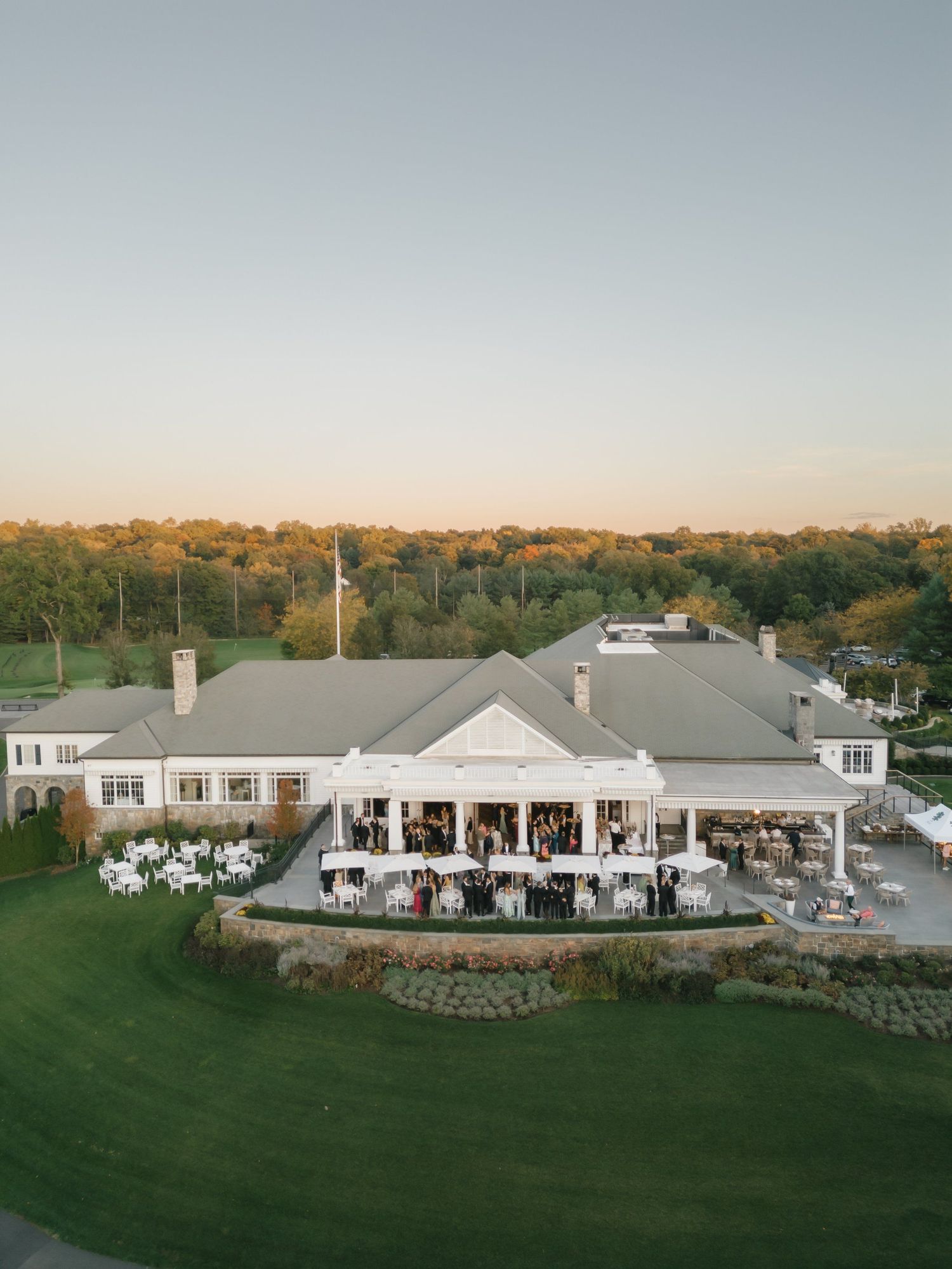 Aerial view of an elegant country club venue with white outdoor dining terrace surrounded by lush green lawns at golden hour.