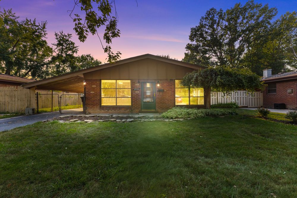 Brick ranch home illuminated at dusk with lush green lawn, mature trees, and wooden fence at twilight.