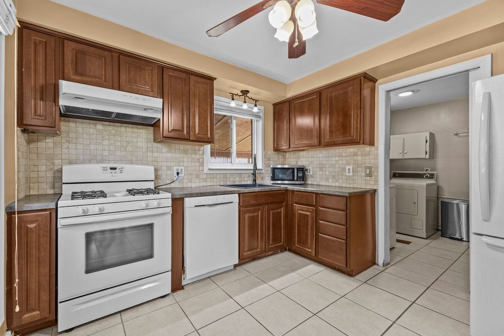 Kitchen with brown cabinetry, tile backsplash, white appliances, tile floor, and ceiling fan with washer visible.