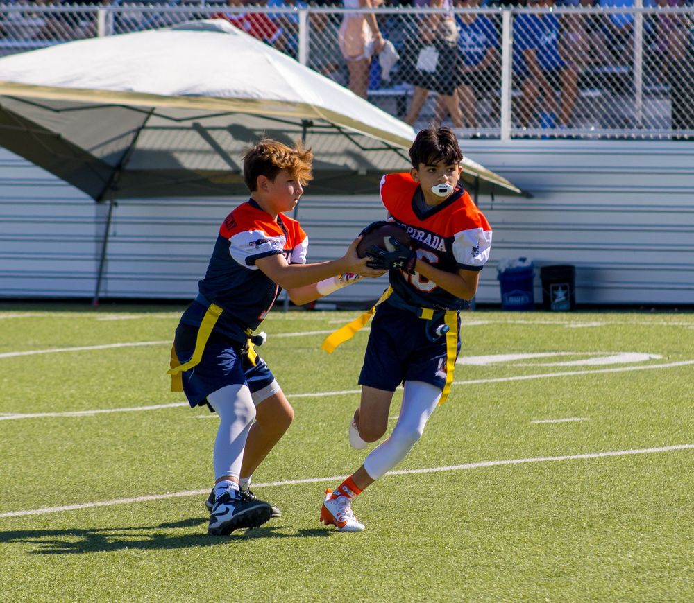 Two youth players compete in a flag football game on a green turf field during a sunny outdoor tournament.