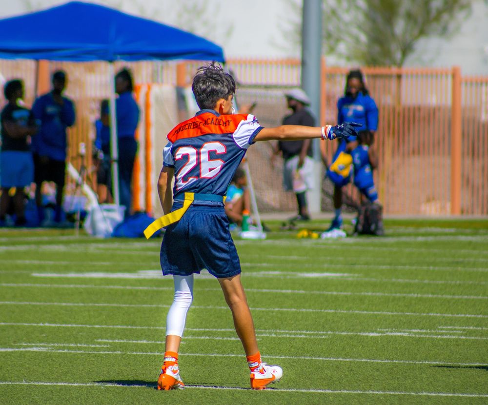 Young flag football player #26 in blue and orange uniform reaches out on green turf field during youth sports game.