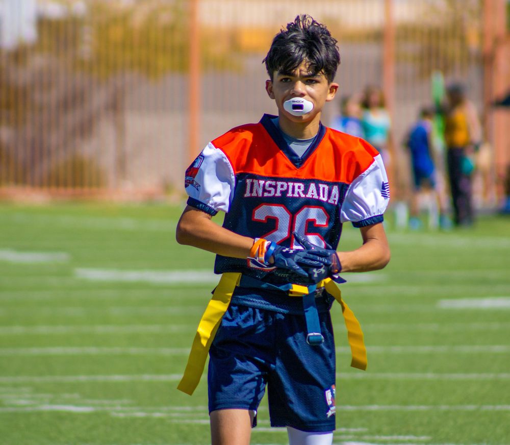 Young football player in Inspirada #26 jersey with mouthguard and flag football belt on green field.