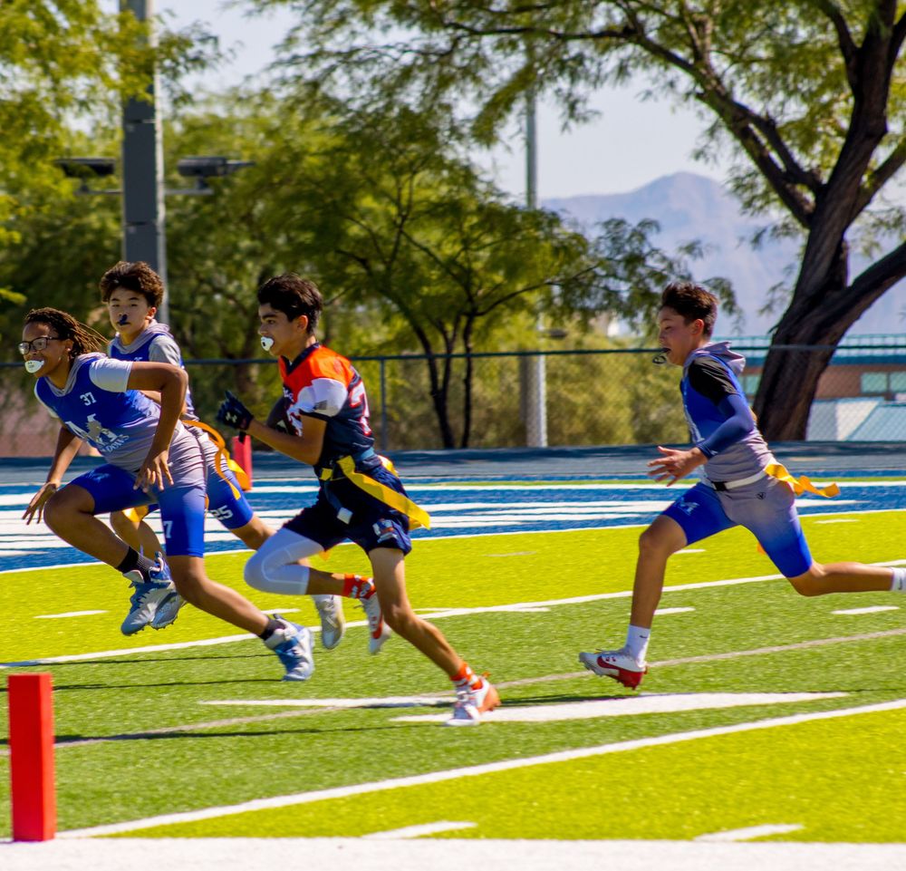 Young boys playing flag football on a blue turf field on a sunny day, running and competing outdoors.