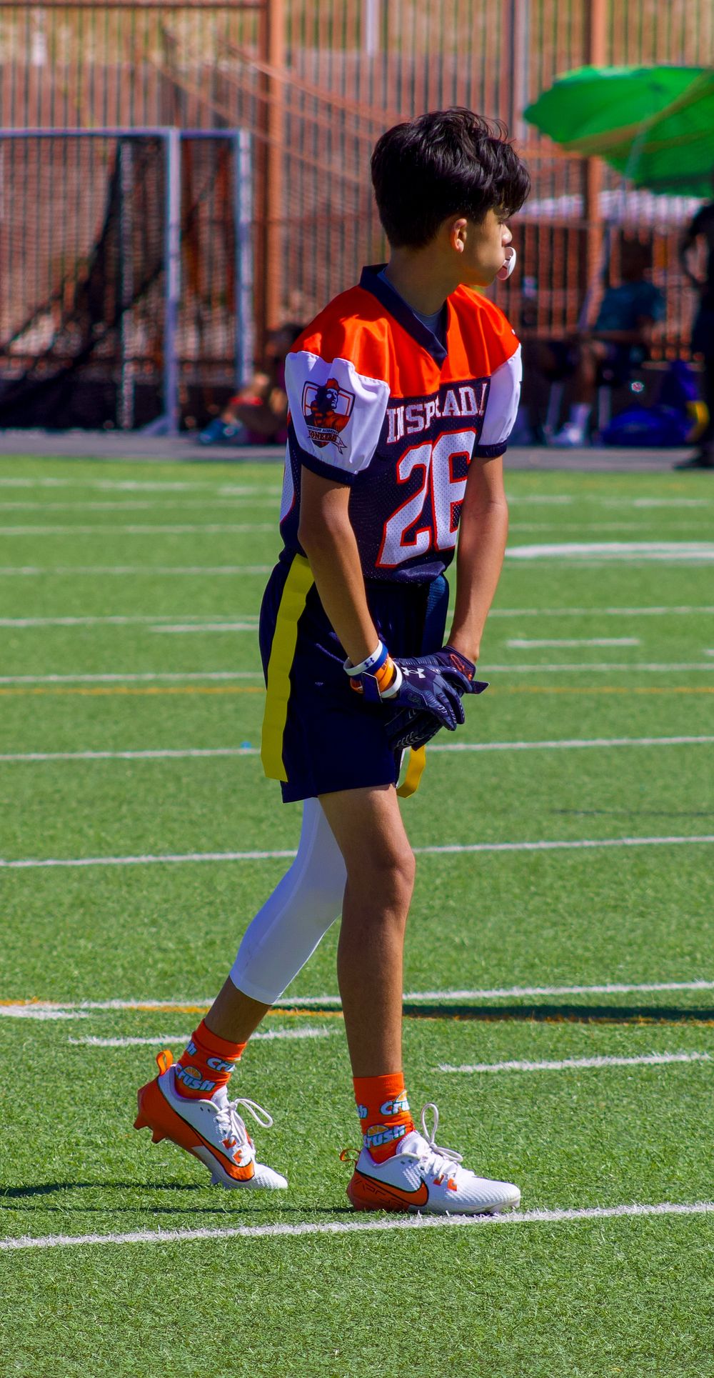 Young football player in orange and navy jersey number 26 stands on green turf field wearing white cleats and orange socks.