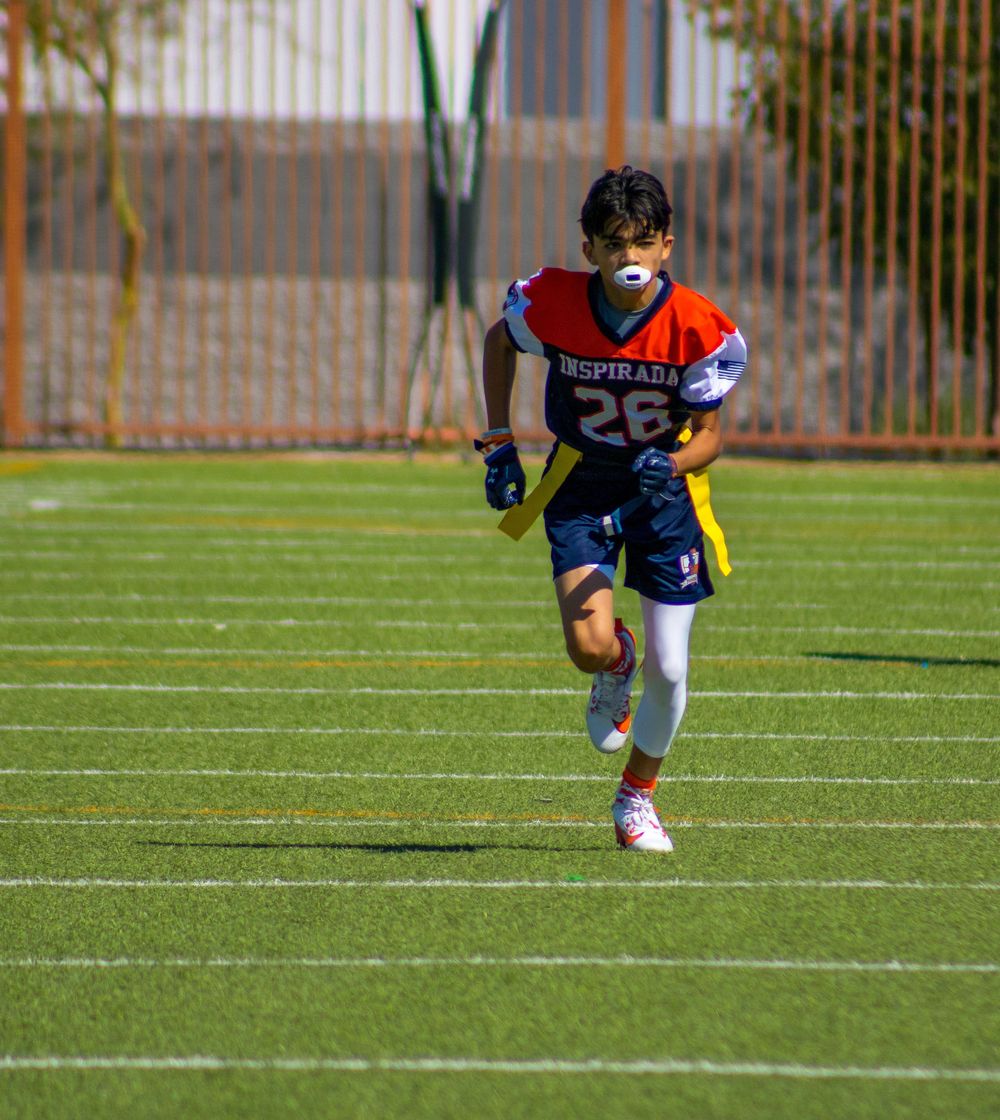Young flag football player #26 in red and blue Desperados jersey runs on green turf field during a sunny game.