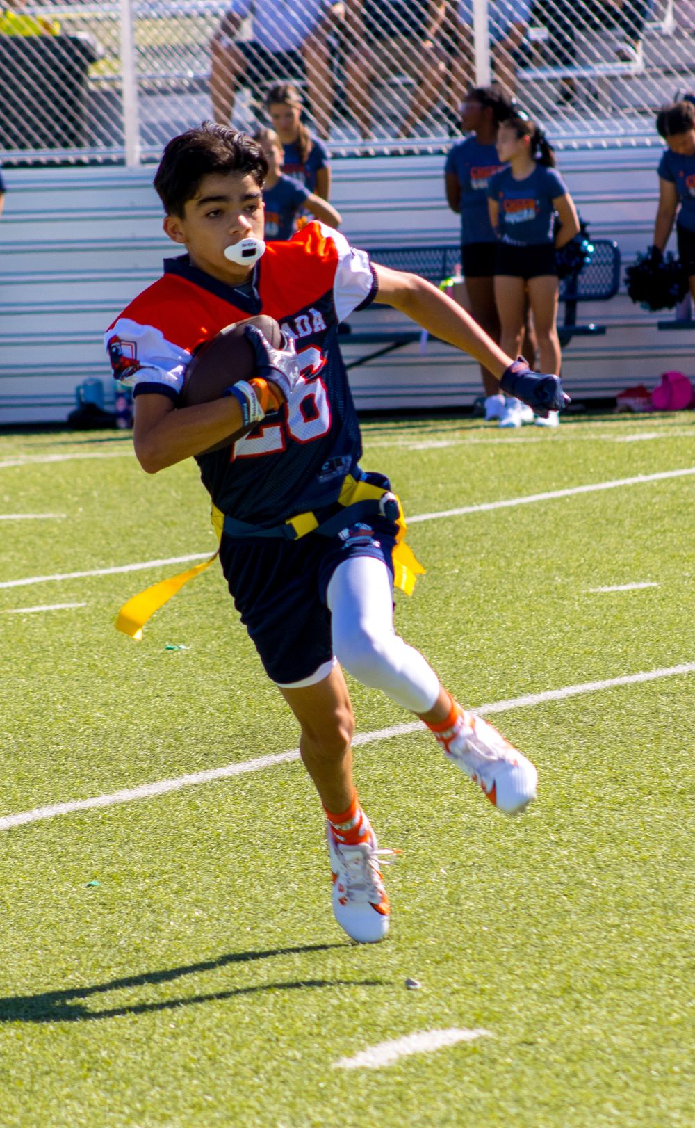 Young flag football player in navy and orange jersey runs with ball during a sunny outdoor game on green turf field.