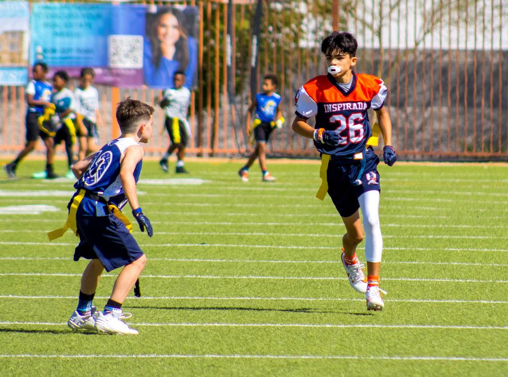 Two youth flag football players compete on a green turf field, player #26 in orange jersey runs past defender.