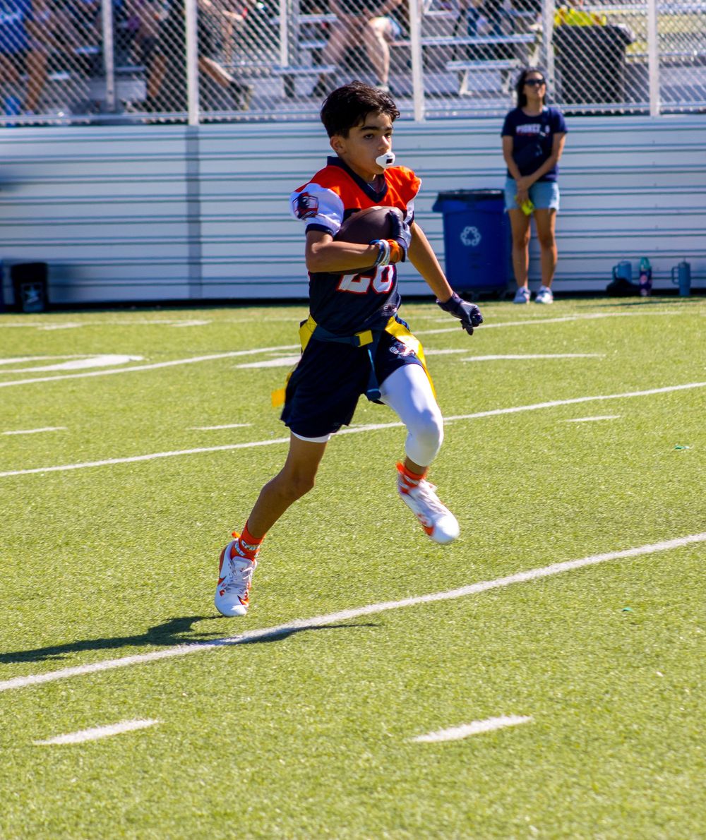 Young football player in navy and orange uniform running on green turf field during practice.