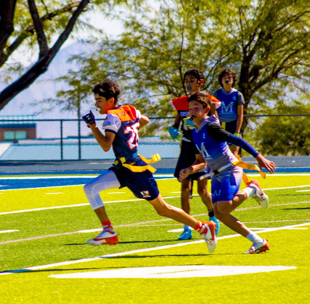 Young athletes compete in a flag football game on a bright green turf field on a sunny day.