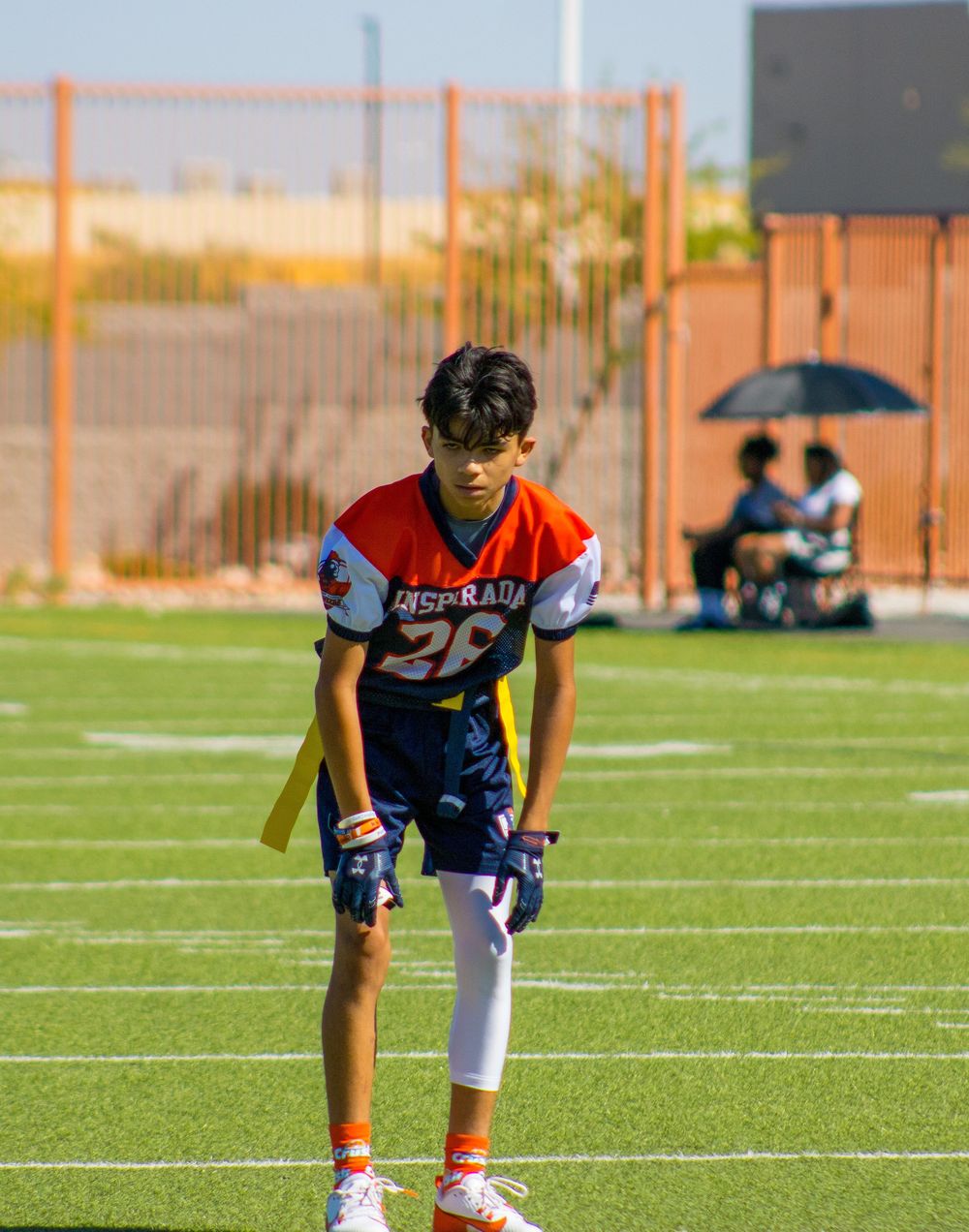 Young flag football player in blue and orange jersey number 28 crouches in ready stance on green turf field.