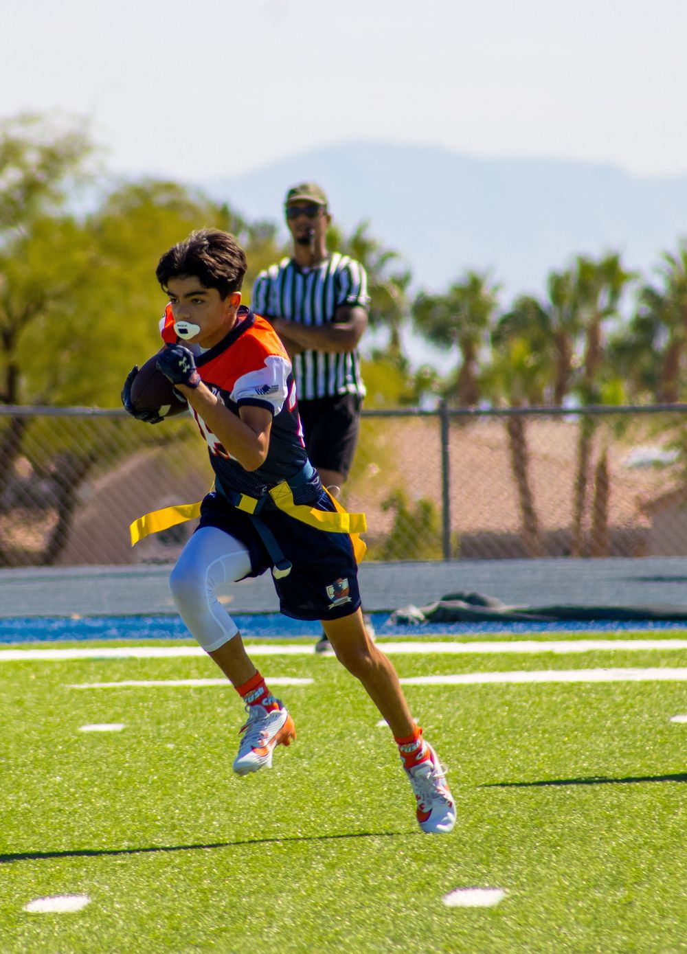 Young athlete runs with football in flag football game on turf field with referee and mountains in background.