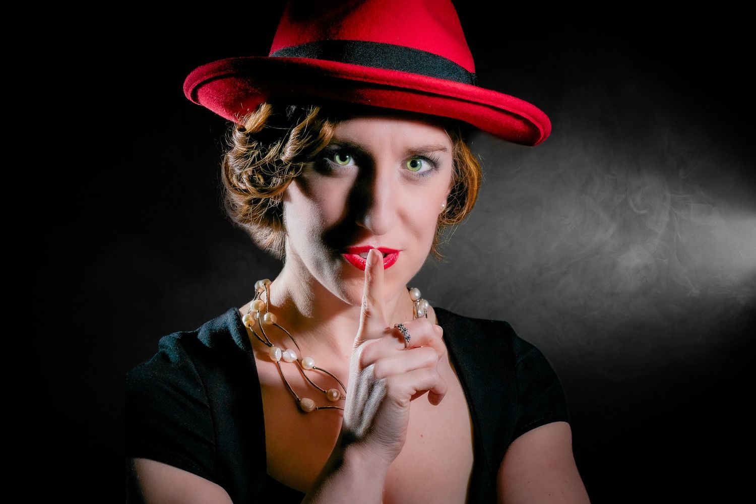 Woman in red hat with finger to lips in shush gesture, wearing pearls and red lipstick on black background.