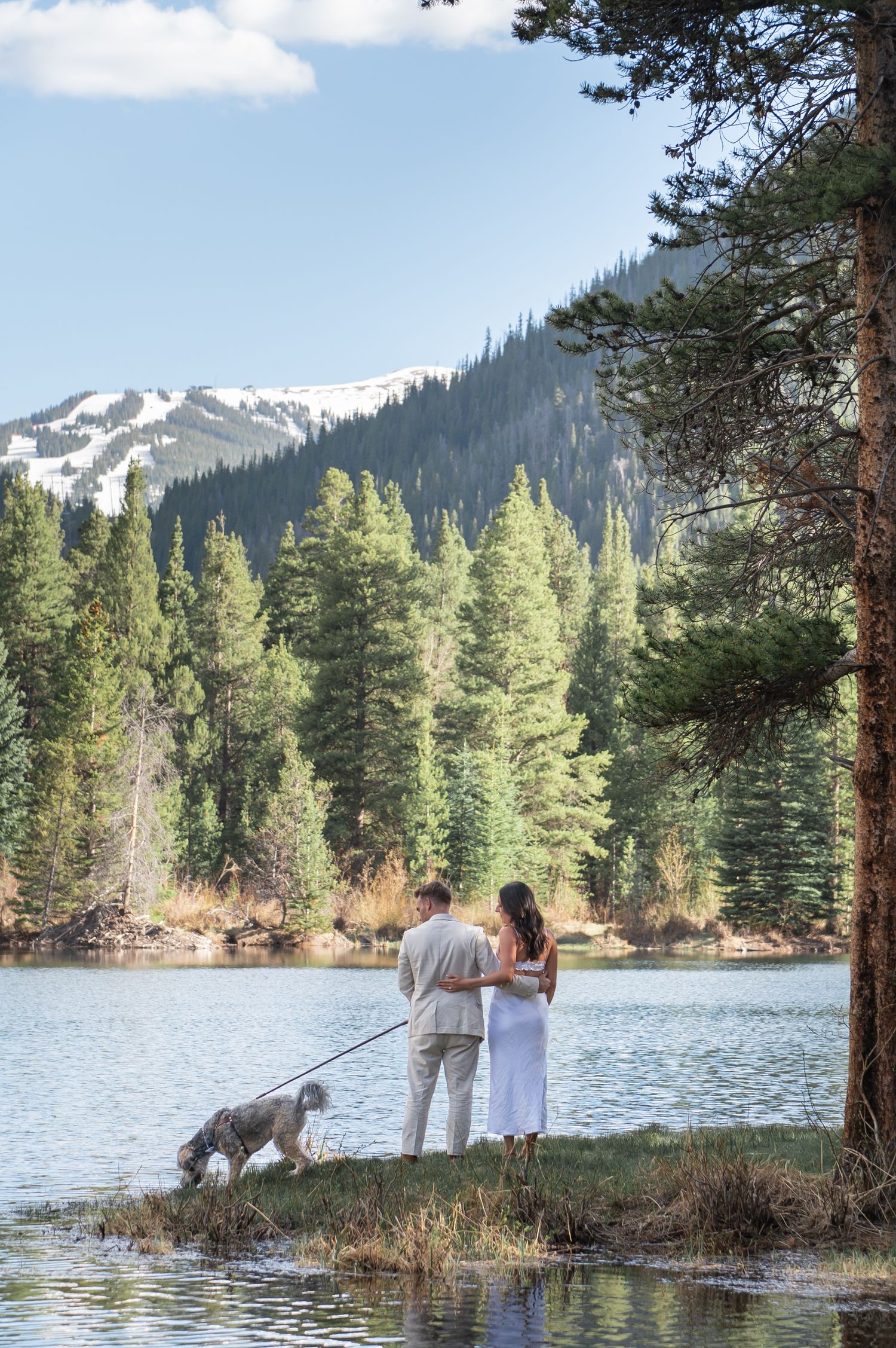 Couple with dog standing by a mountain lake surrounded by pine trees and snow-capped peaks on a sunny day Officer's Gulch elopement
