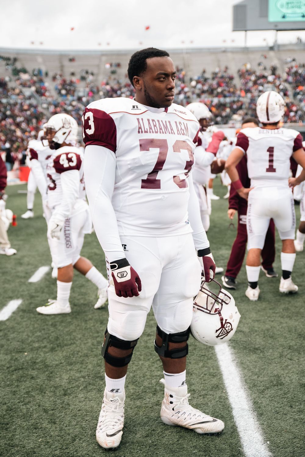 Alabama A&M football player #73 stands on sideline in white uniform holding helmet during a college game.