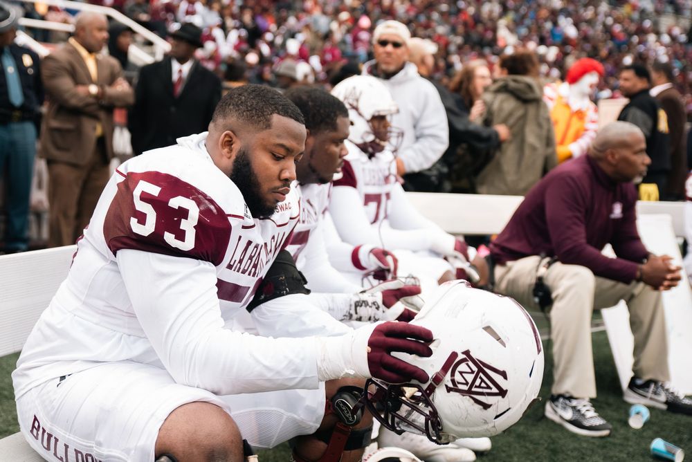 Football player #53 in maroon and white uniform sits on sideline bench with helmet, looking downward during game.