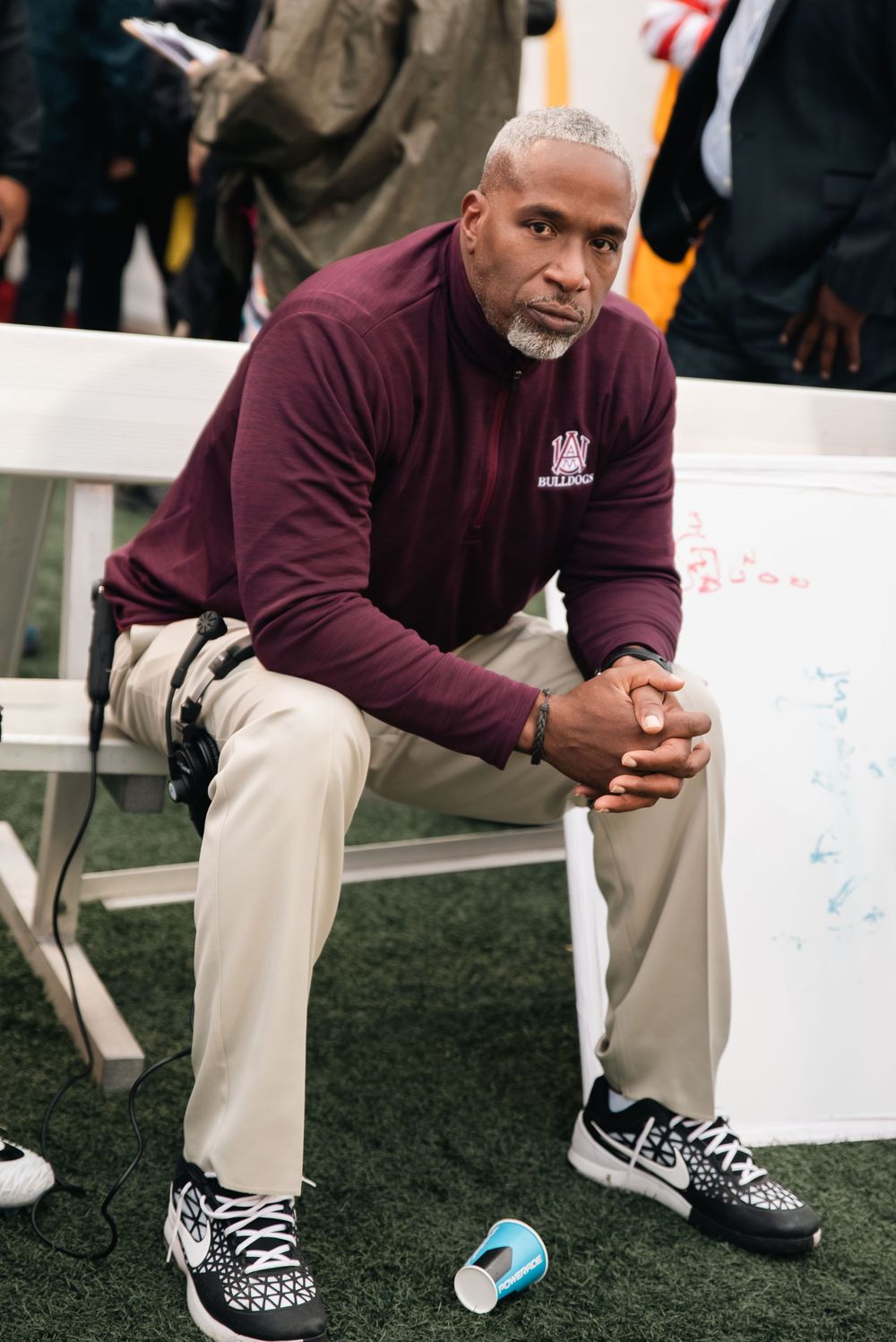 A football coach in a maroon Bulldogs pullover sits on a bench, looking focused during a game.