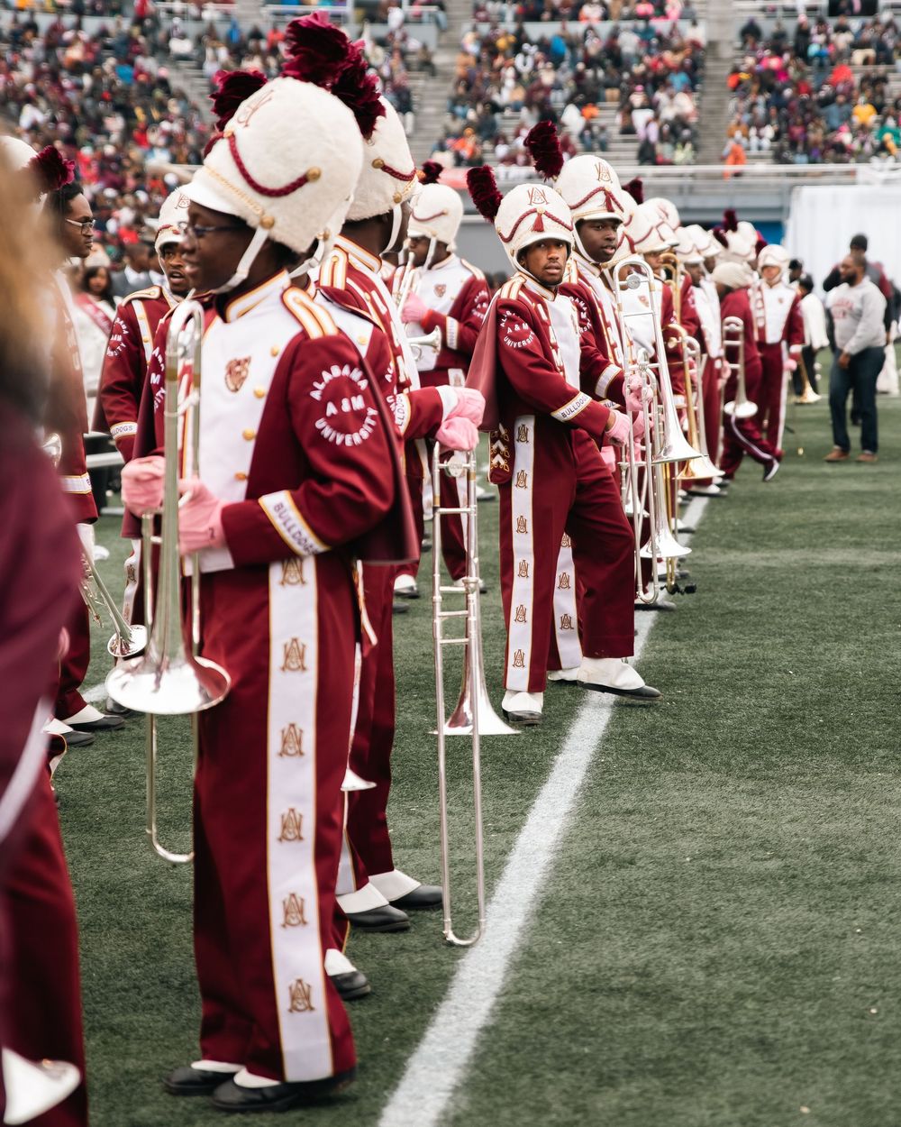 Marching band members in red and white uniforms with plumed hats perform on a football field during a game.