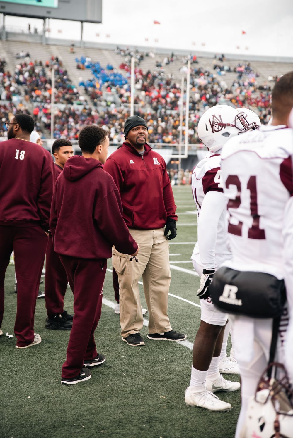 Football coach in maroon tracksuit talks to players on sideline during a packed stadium game day.