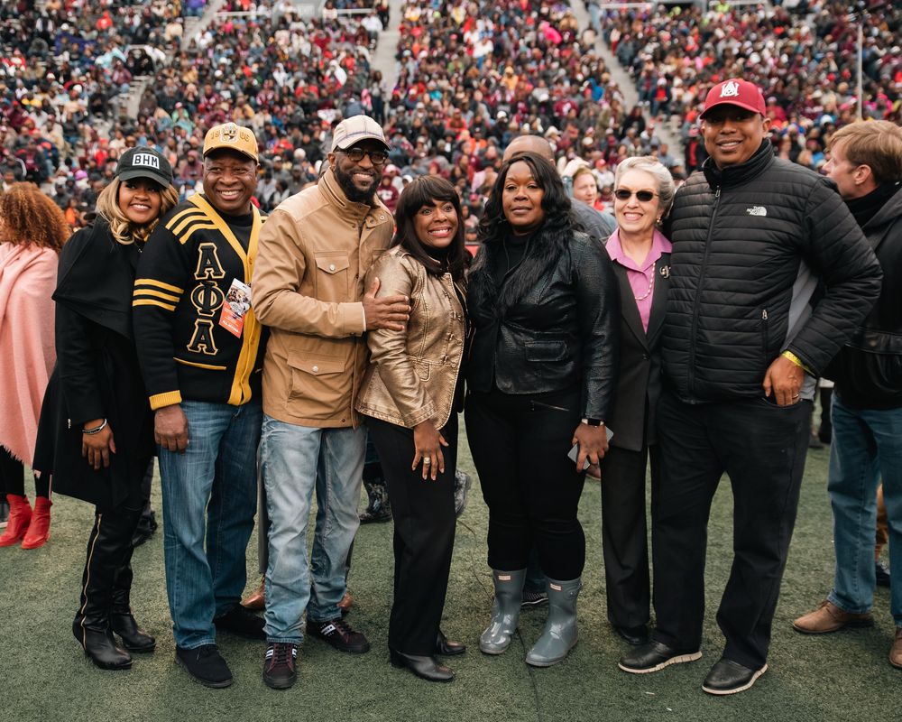 Group of people posing together at a football stadium during a game, smiling for a photo on the sidelines.