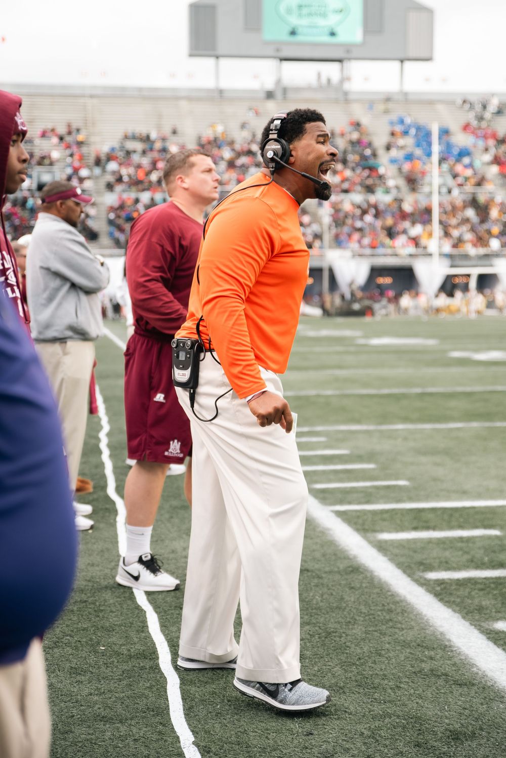 Football coach in orange jacket with headset yelling on sideline during a college football game at a packed stadium.